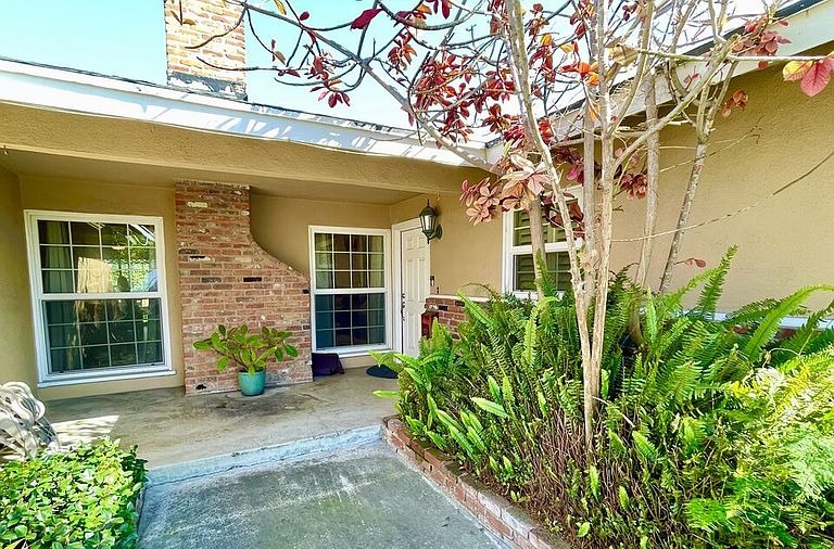 Side patio at 16622 Tiber Ln with brick chimney detail, white windowsill, and ferns framing a covered porch area