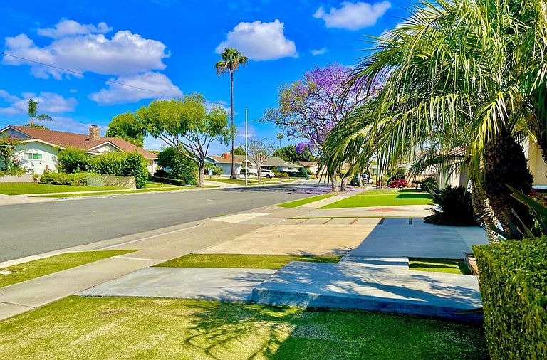 Quiet residential street view from 16622 Tiber Ln looking down a tree-lined Huntington Beach block with a tall palm and a flowering jacaranda overhead