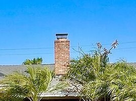 Tight architectural detail of the brick chimney at 16622 Tiber Ln, rising from the original 1960 ranch roofline against a clear blue sky with palm fronds framing the right edge