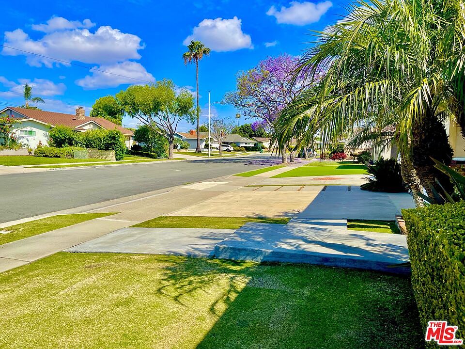 Looking down Tiber Lane toward the home from the curb, the street empty and tree-lined with a jacaranda in bloom against a blue sky